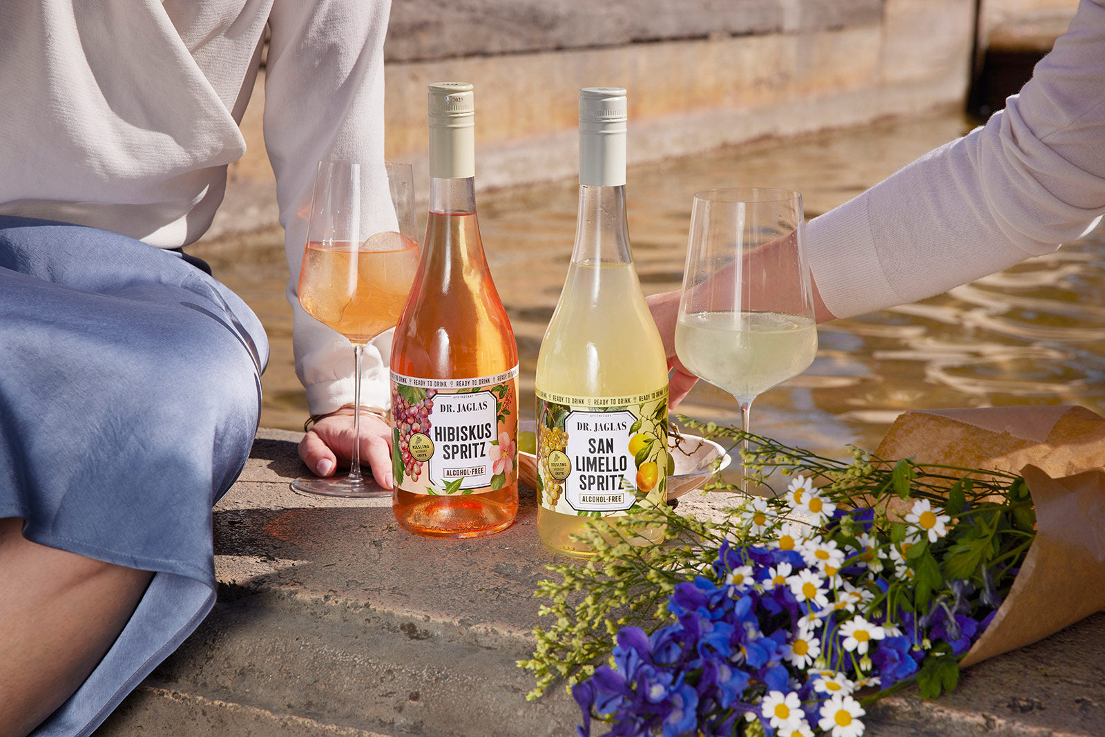 Zwei Personen entspannen am Wasser mit dr-jaglas Hibiskus Spritz - 5 + 1 Bundle, genießen Gläser des veganen, kalorienarmen Getränks und Blumensträuße aus blauen und weißen Blüten an ihrer Seite.