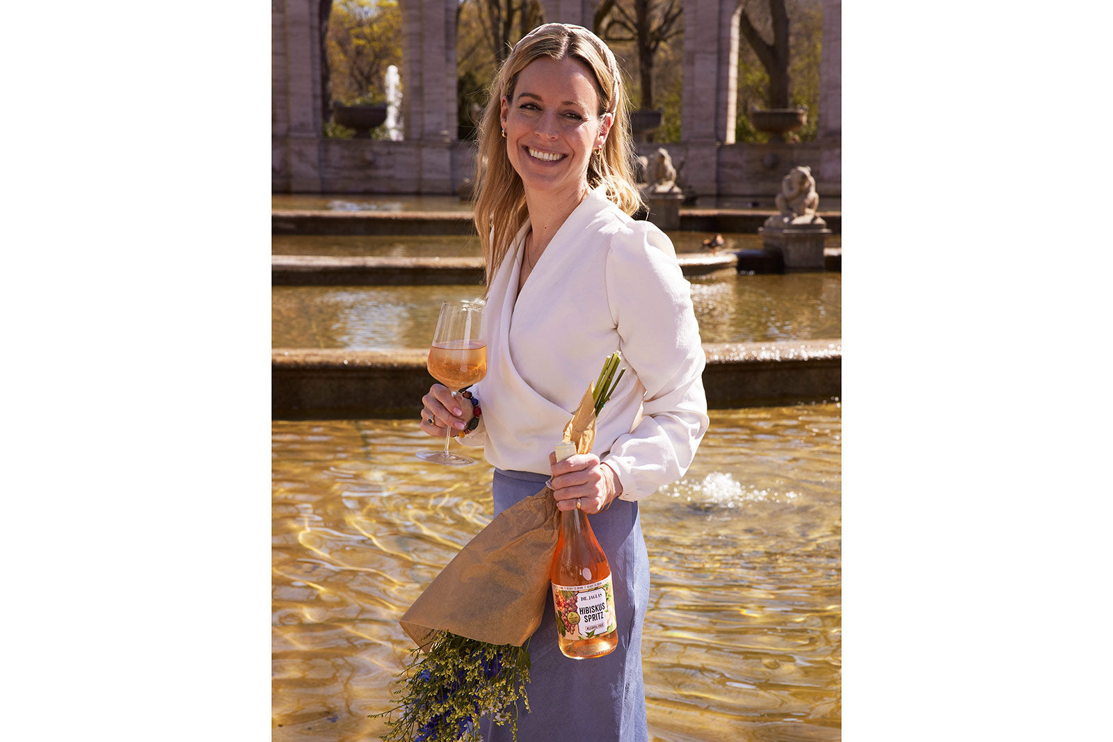 Eine lächelnde Frau steht an einem Brunnen und hält ein Glas dr-jaglas Hibiskus Spritz und eine Flasche aus dem Hibiskus Spritz - 5 + 1 Bundle zusammen mit einem Blumenstrauß.