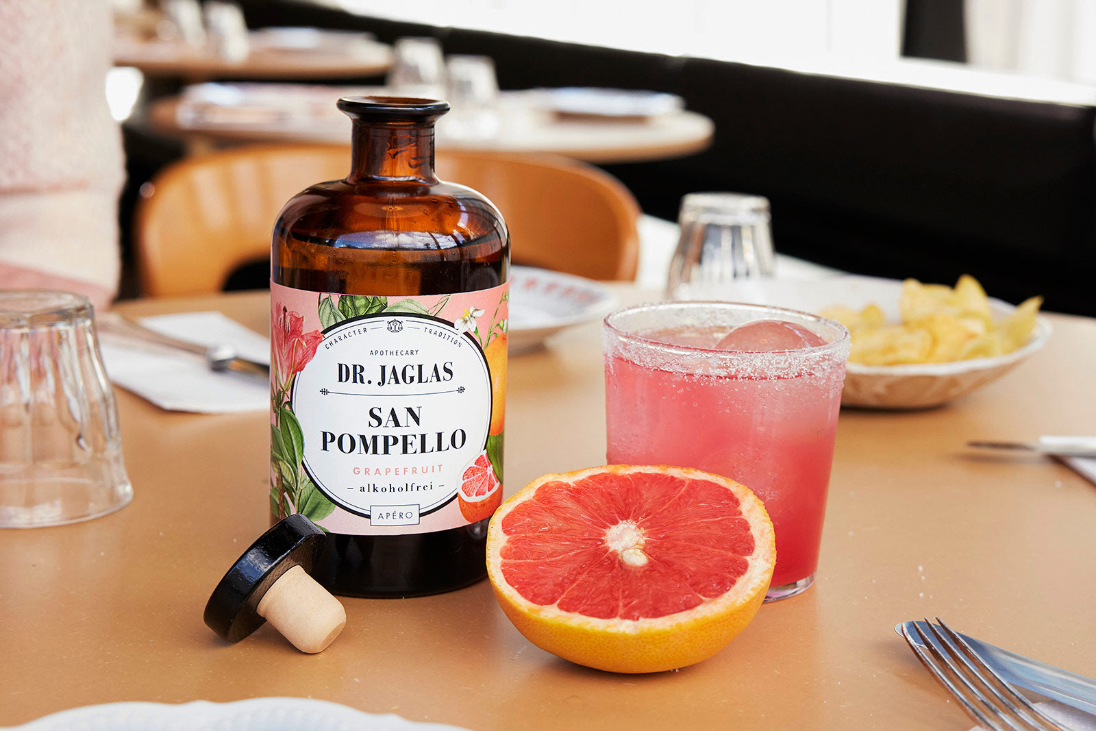 Drei Flaschen dr-jaglas San Pompello im Bundle, ein Glas rosa Aperitif und eine halbe Grapefruit - perfekt für Ihren nächsten Sommerdrink oder Ihr Paloma-Rezept.
