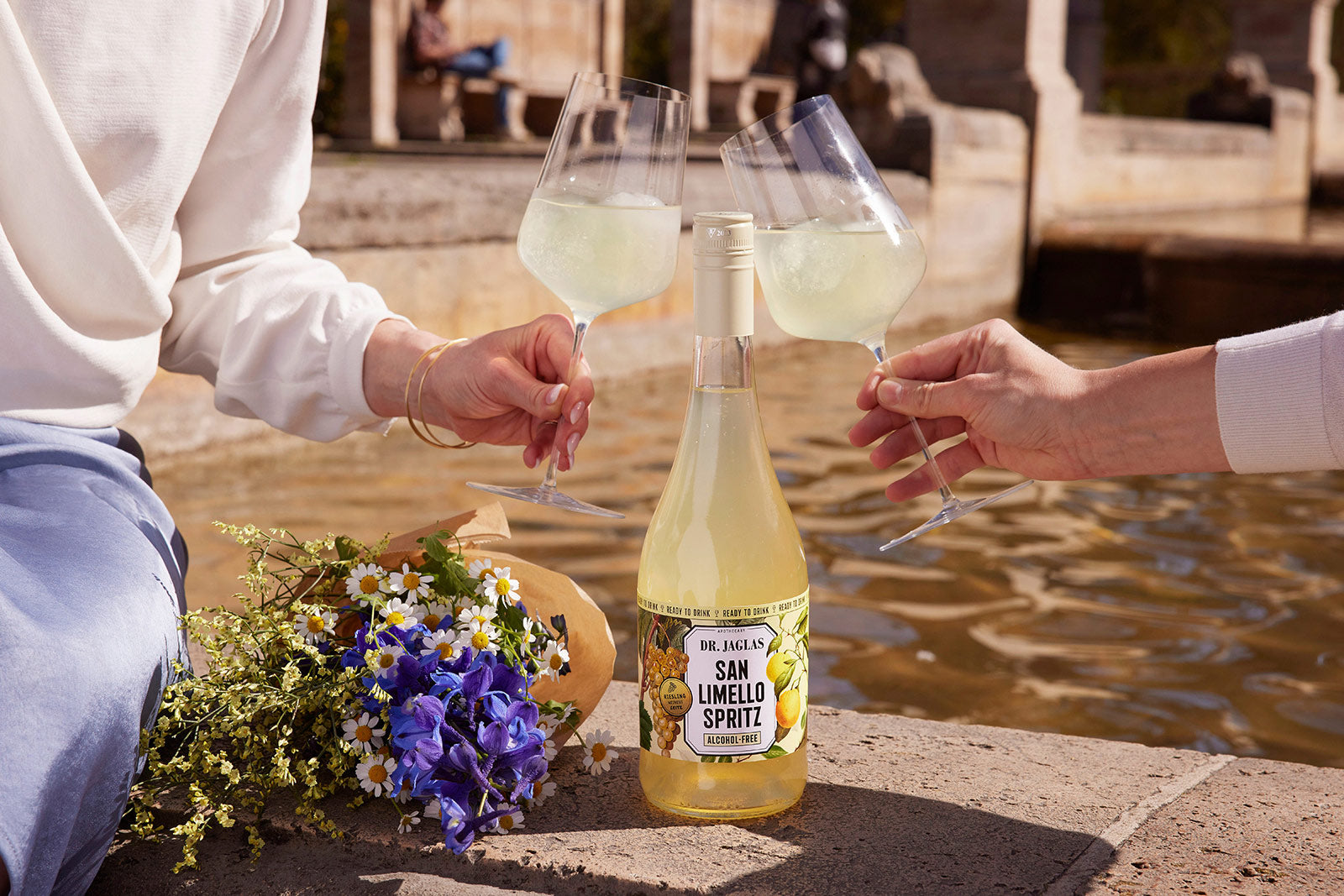 Drei Personen stoßen mit dr-jaglas Kennenlern-Bundle an einem Brunnen an, mit einer Flasche und Blumen in der Nähe - und genießen einen veganen, alkoholfreien Moment zusammen.