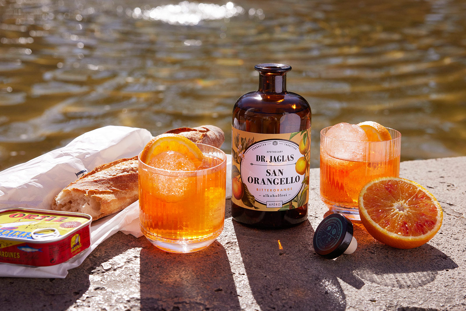 Zwei Orangencocktails, eine Flasche dr-jaglas San Orangello, Brot, Sardinen und eine Orange liegen am Wasser im Sonnenlicht.