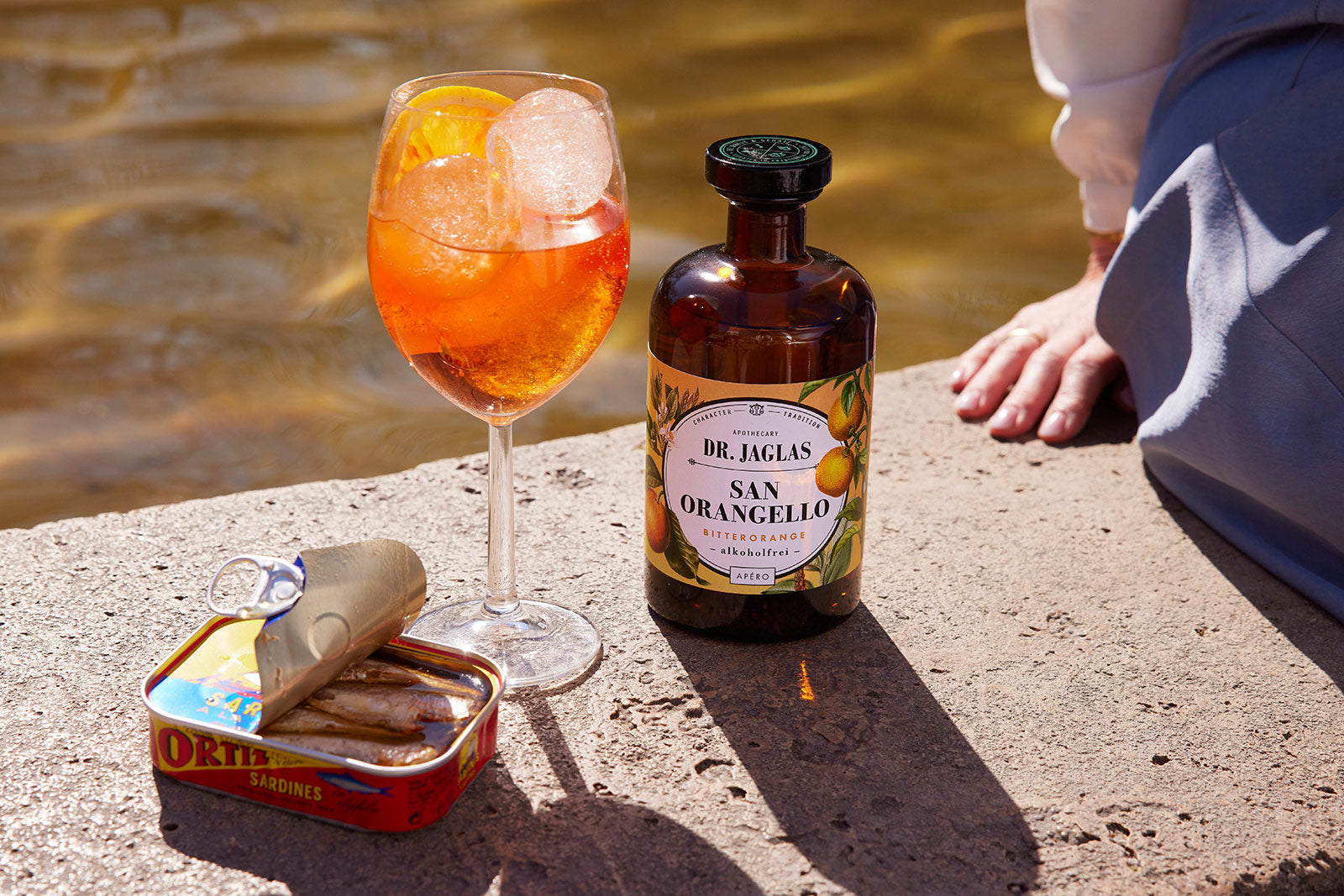 Ein Glas Orangencocktail, eine Flasche dr-jaglas San Orangello und eine geöffnete Sardinendose stehen auf einem Ufervorsprung.