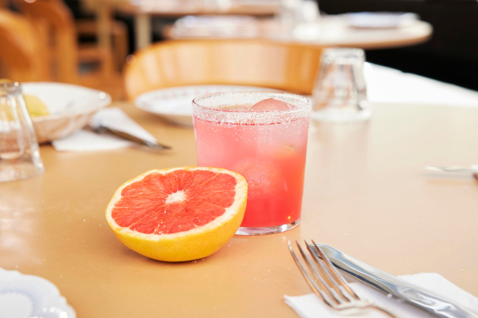 Ein gekühltes Glas dr-jaglas 3x San Pompello im Bundle, das perfekte Sommergetränk oder ein rosafarbener Aperitif, steht neben einer halben Grapefruit auf einem Restauranttisch.