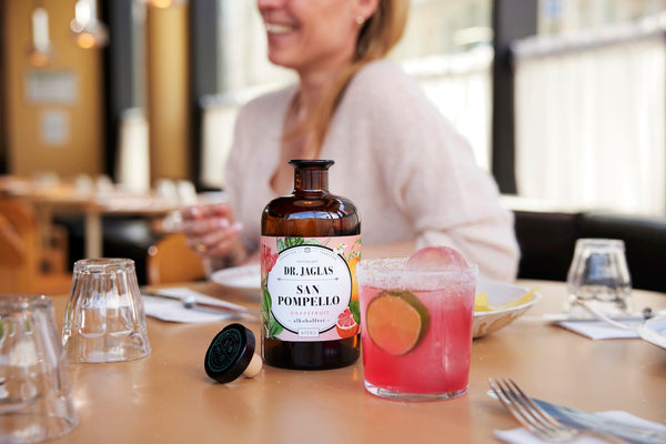 Eine Flasche aus dem dr-jaglas Kennenlern-Bundle und ein rosafarbener Hibiskus-Spritz stehen auf einem Tisch mit einer lÀchelnden Frau im Hintergrund, die den alkoholfreien und veganen Genuss prÀsentiert.